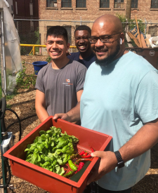 Picture of three men outdoors on a sunny day, smiling at the camera. the man in the foreground is holding a red box with plant seedlings in it. A red bridk building can be seen in the background. A garden hoop house is seen at left. 